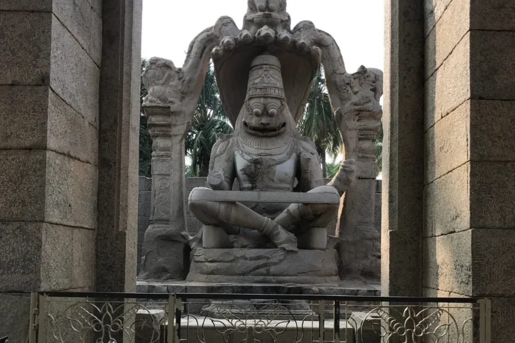 The massive stone monolith sculpture of Lord Narasimha, the half-man half-lion avatar of Vishnu, sitting cross-legged under a multi-headed serpent canopy at the Shri Lakshmi Narasimha Temple ruins in Hampi, framed by large stone entry pillars.