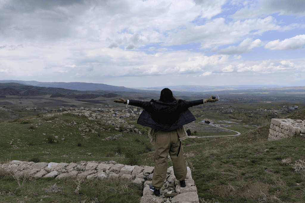 Sumana with her arms outstretched, standing on the high rocky plateau of Büyükkale overlooking a wide valley.