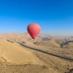 A red hot air balloon soaring above sandy desert hills and mountains in Luxor under a clear sky.