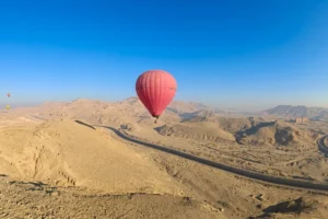 A red hot air balloon soaring above sandy desert hills and mountains in Luxor under a clear sky.