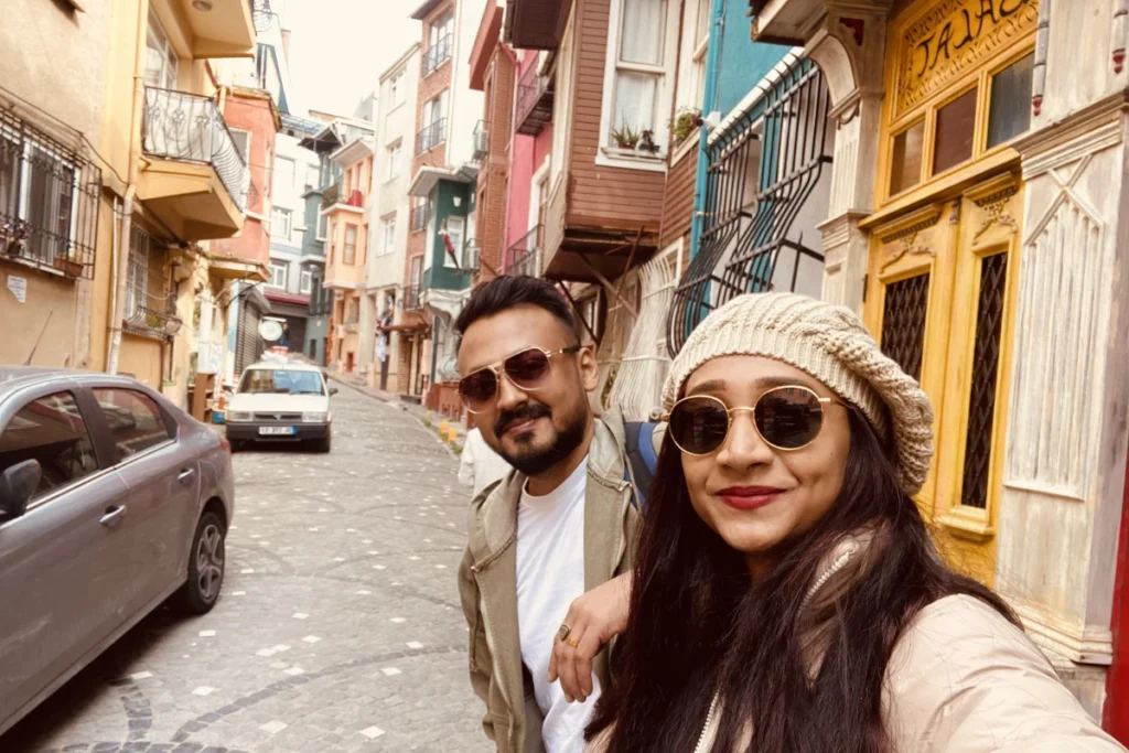 Istanbul- Balat-2 Akash & Sumana smiles for a close-up selfie while wearing sunglasses and winter beanies, standing on a cobblestone road with colorful buildings behind them.