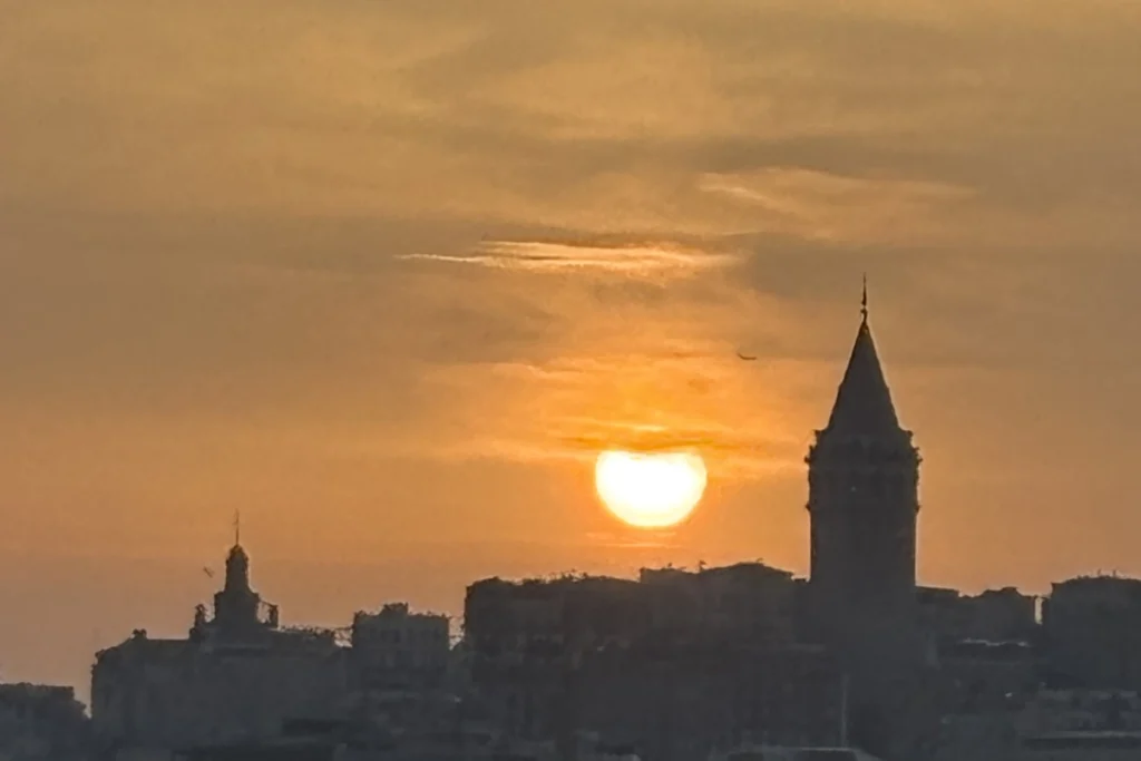 Istanbul- Cruise - Sunset A silhouette of the distant Galata Tower cutting into a fiery, dark orange sunset sky over the water.