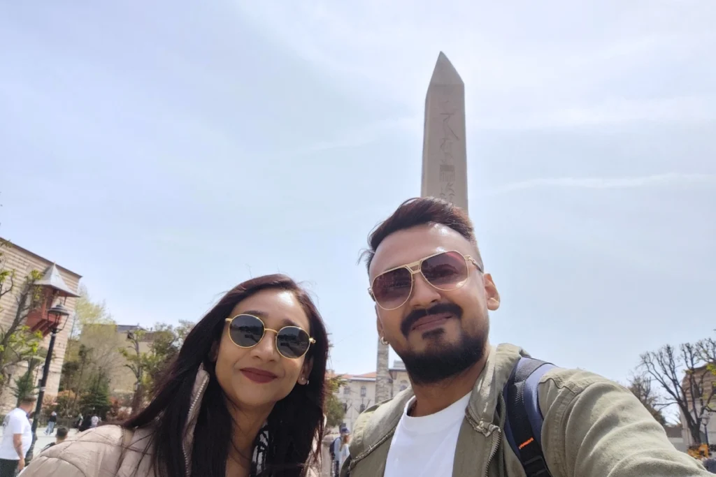 Istanbul- Egyptian oblique Akash & Sumana taking a close-up selfie in a public square with the towering ancient stone Egyptian Obelisk in Istanbul standing directly behind them.