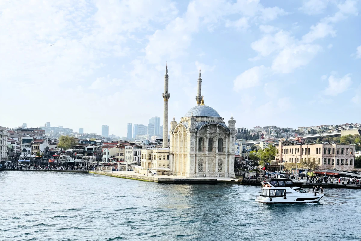 A wide scenic view of Istanbul showing the grand white Ortaköy Mosque with its distinct domes and minarets perched on the waterfront against a sprawling cityscape.