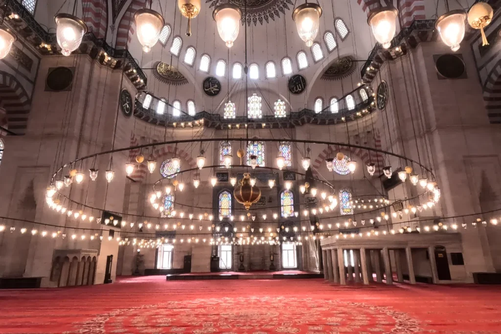 Istanbul- Sulemaniye Mosque An internal perspective of the vast Süleymaniye Mosque showing massive low-hanging circular chandeliers illuminating the patterned carpet below.
