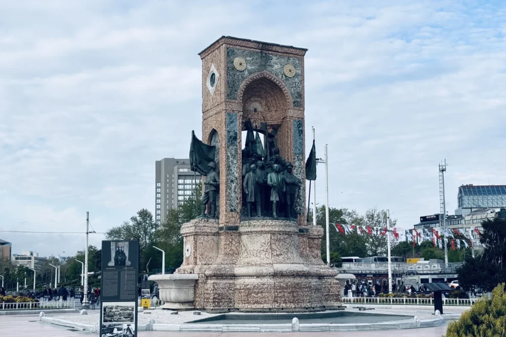 Istanbul- Taksim square- Republic Monument A high stone monument featuring detailed bronze sculptures of historical figures standing in the center of an open public square.