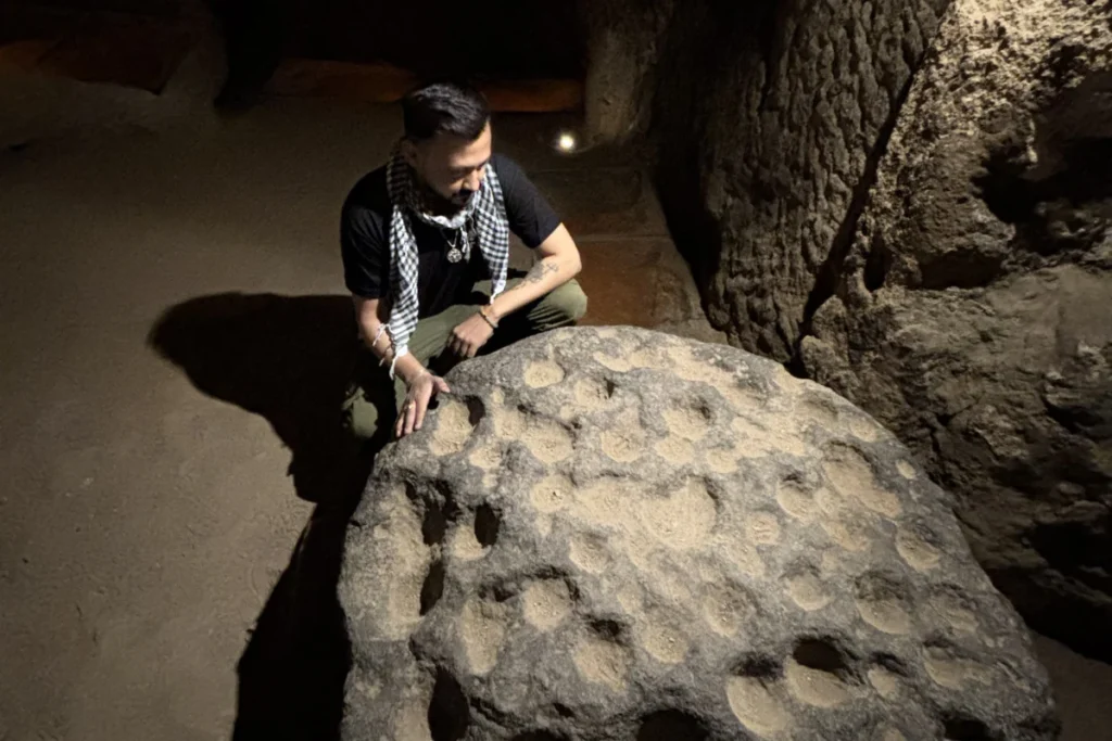 Akash stands inside a stone room touching a massive, circular millstone-style rolling door once used to block entrance tunnels.
