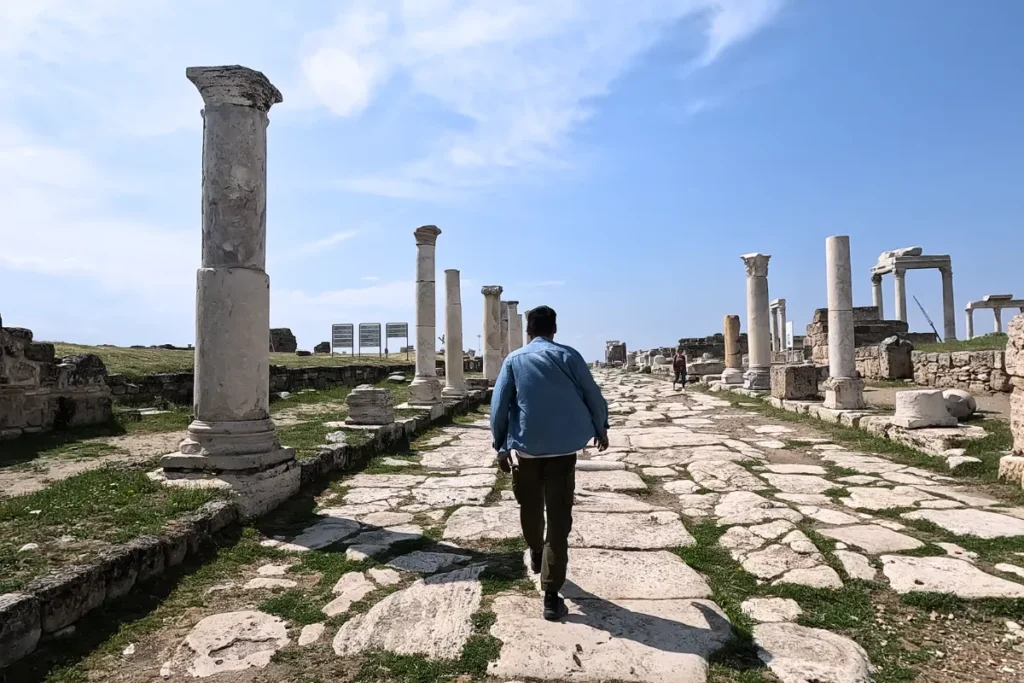 Akash seen from behind walking down a dirt path flanked by standing ancient Roman stone columns and ruins.