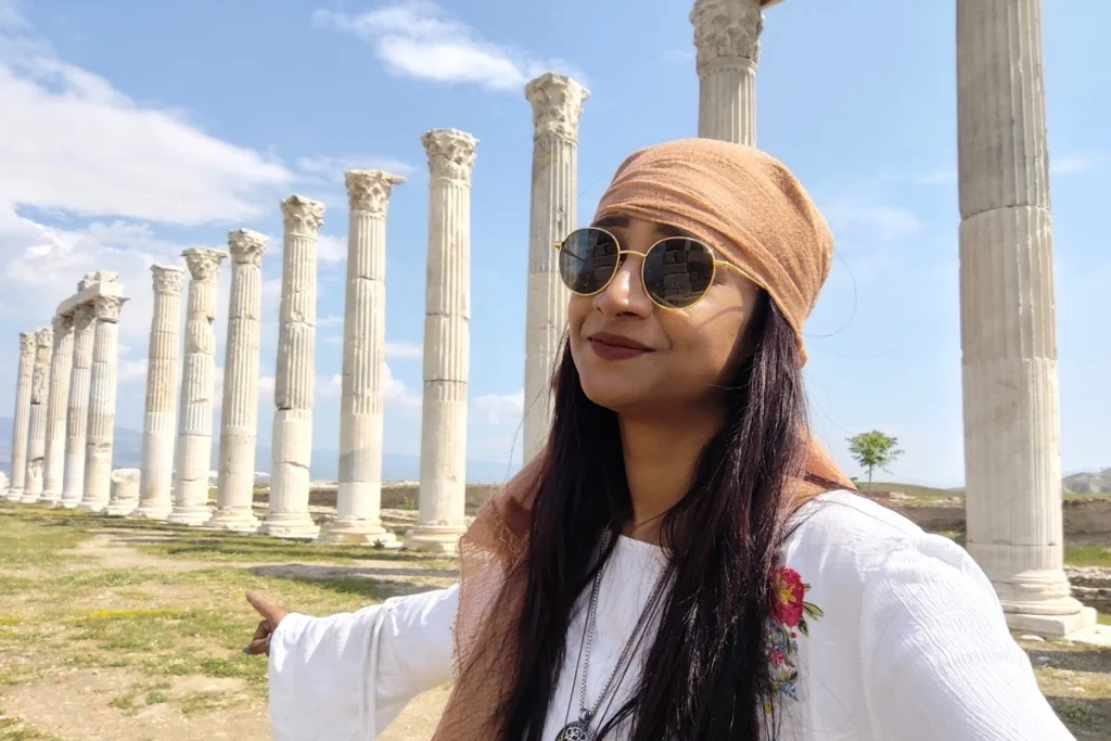 Sumana wearing a white headscarf and sunglasses looking off into the distance amidst standing ancient Roman columns.