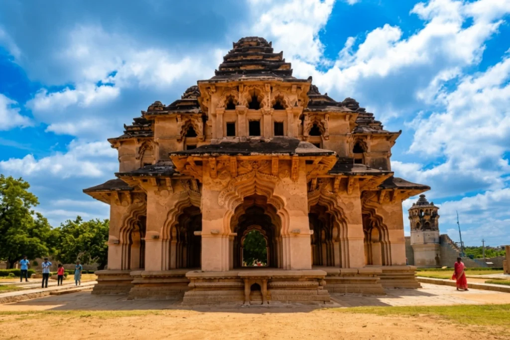 A two-story symmetrical stone pavilion with intricate arches and dome roofs known as the Lotus Mahal, surrounded by a lawn.