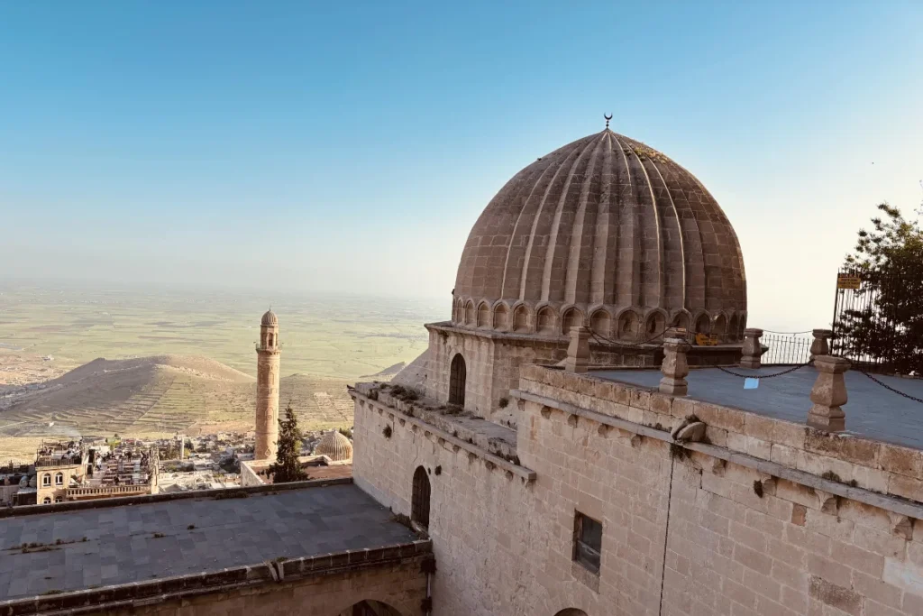 A wide view showing the stone domes of the Zinciriye Madrasa overlooking the vast, flat Mesopotamian plain under a hazy sky.