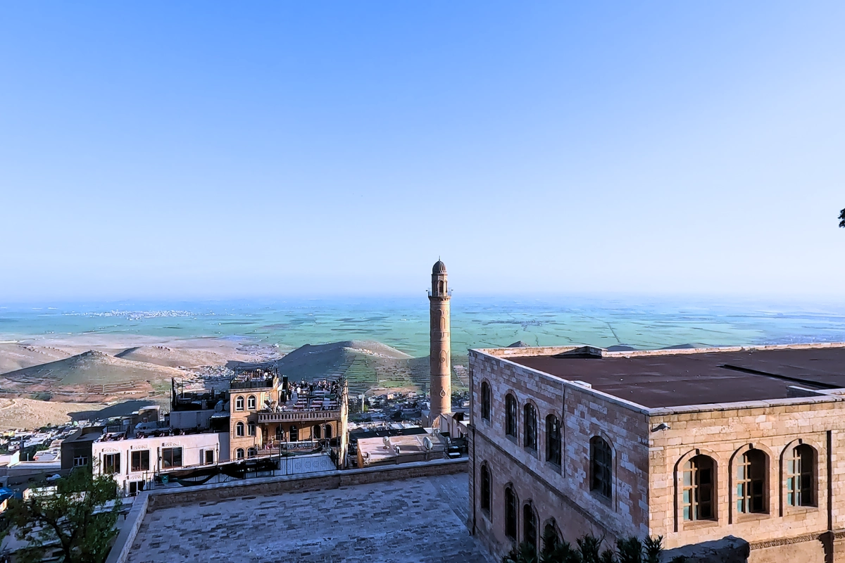 A wide panoramic banner image displaying the ancient stone architecture of Mardin, featuring a central tall minaret tower and traditional stone buildings under a bright blue sky, with a expansive landscape stretching out into the distance.