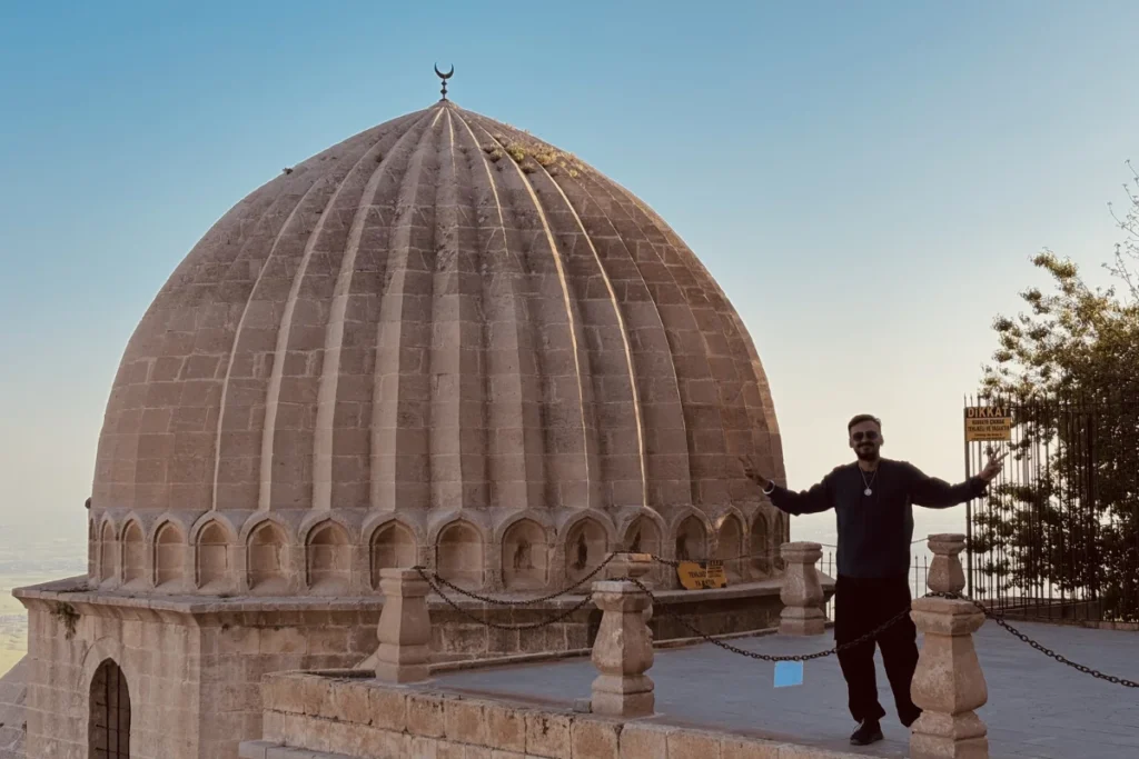 A full-length shot of Akash standing on a stone terrace next to a large, historically detailed fluted stone dome topped with a crescent moon symbol at Zinciriye Madrasa