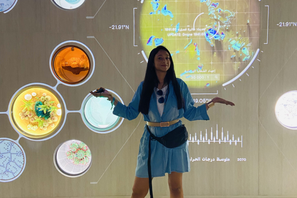Ssumana standing in front of a curved, interactive digital wall displaying glowing blue planetary diagrams and informational infographics.