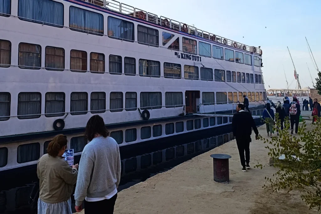 Passengers standing on a concrete pier next to a large, multi-deck white Nile cruise ship.