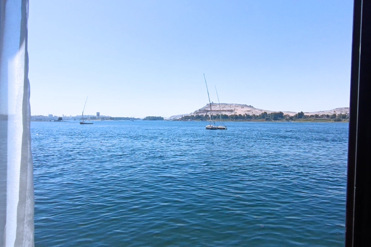 A wide panoramic landscape showing calm blue waters of the Nile River with small boats docked near sandy hills under a clear sky.