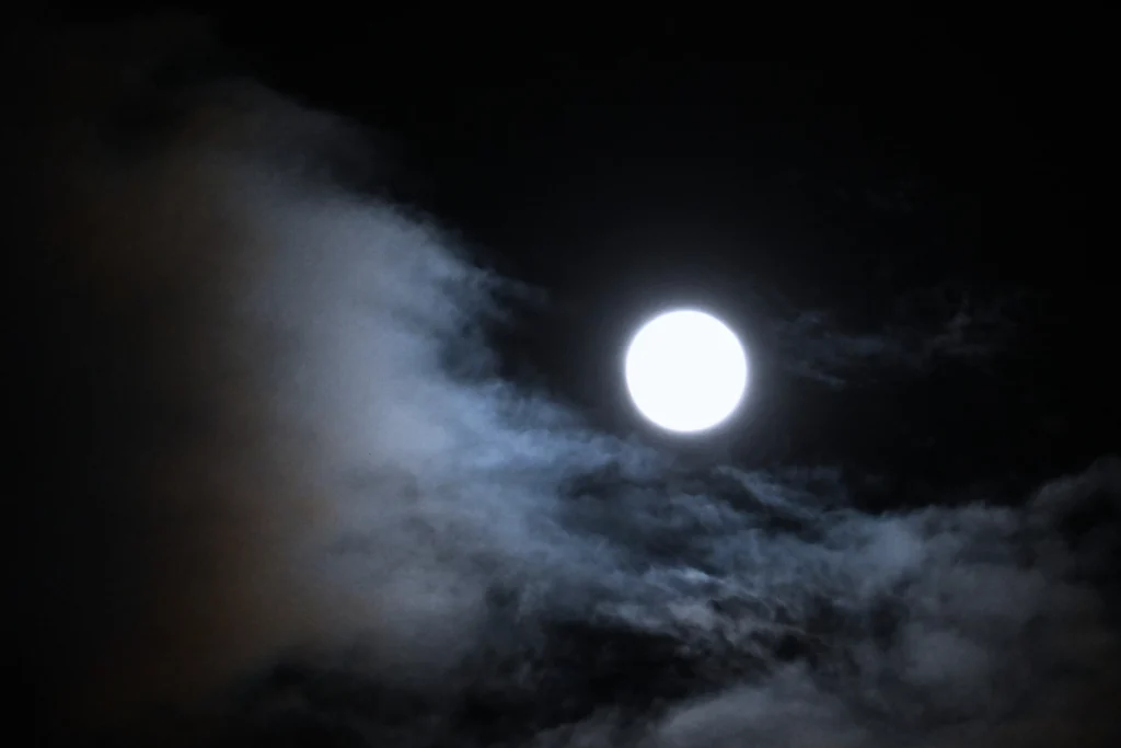 A dramatic night view of a brilliant, glowing white full moon shining brightly through dark, wispy blue and grey clouds in a pitch-black night sky.