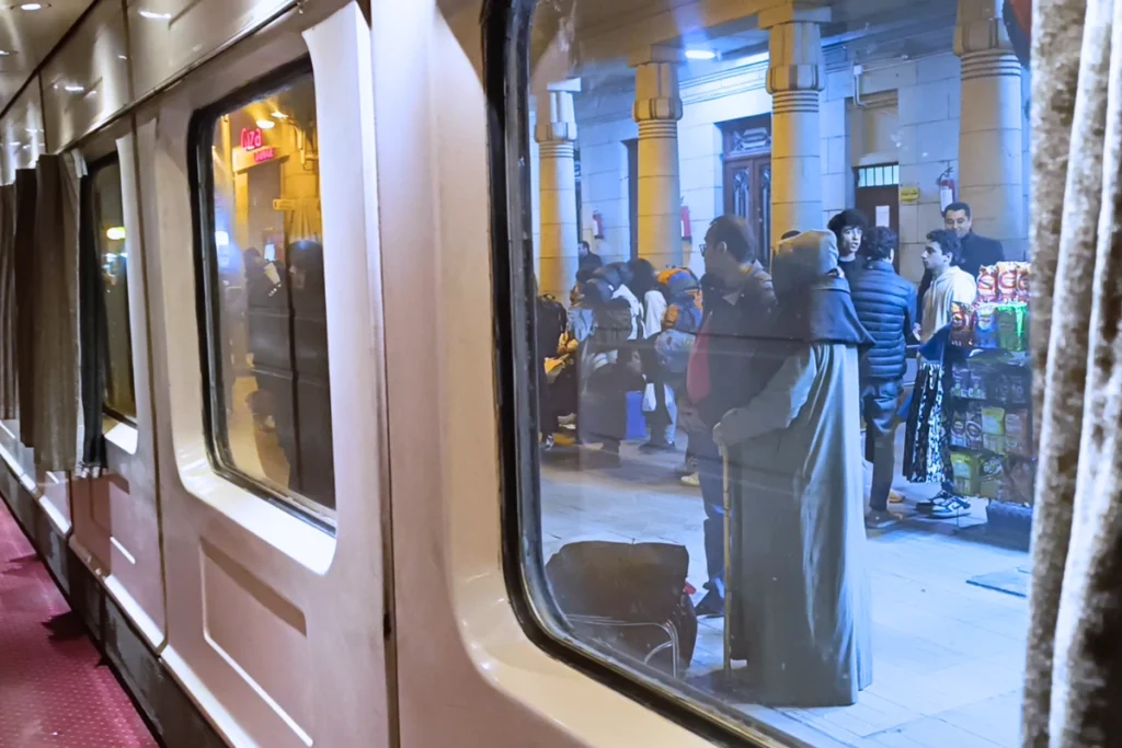 The side of a moving train reflecting the bright lights of a city platform as it departs into the dark night.
