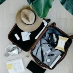 A high-angle flat lay shot of a small black carry-on suitcase packed with folded blue shirts, a sun hat, a pack of wet wipes, travel containers, and white sneakers neatly arranged around it.