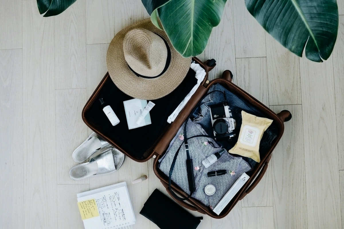 A high-angle flat lay shot of a small black carry-on suitcase packed with folded blue shirts, a sun hat, a pack of wet wipes, travel containers, and white sneakers neatly arranged around it.