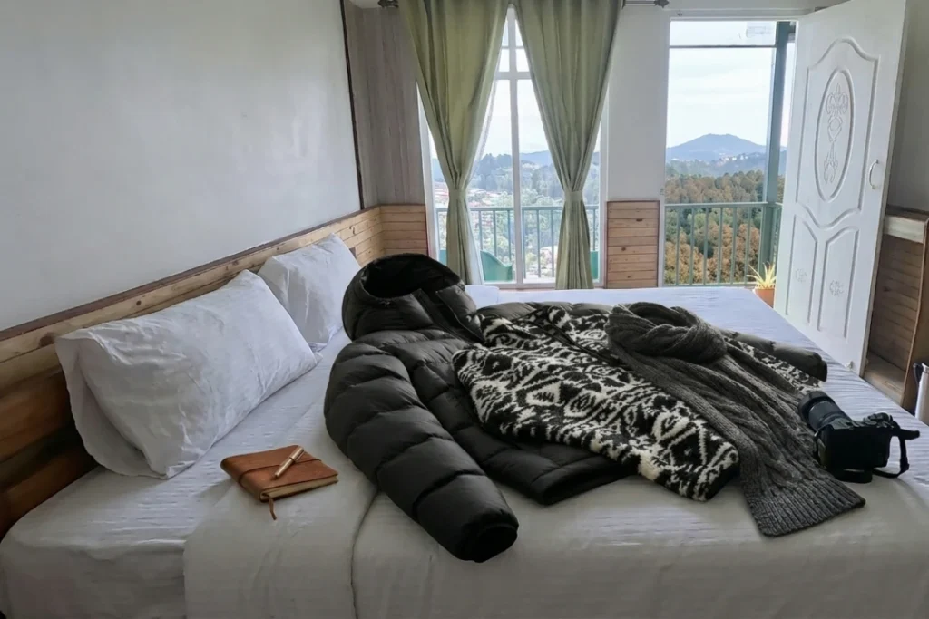 A dark blue winter jacket and multiple pieces of patterned clothing laid flat across a hotel bed with white sheets next to a window.