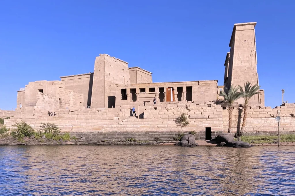 A scenic view across the water showing the ancient stone structures and pylons of the Philae Temple complex on an island.