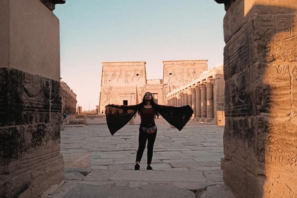 Sumana wearing a dark shirt stands on a stone platform with his arms outstretched in front of an ancient Egyptian temple wall adorned with hieroglyphics.