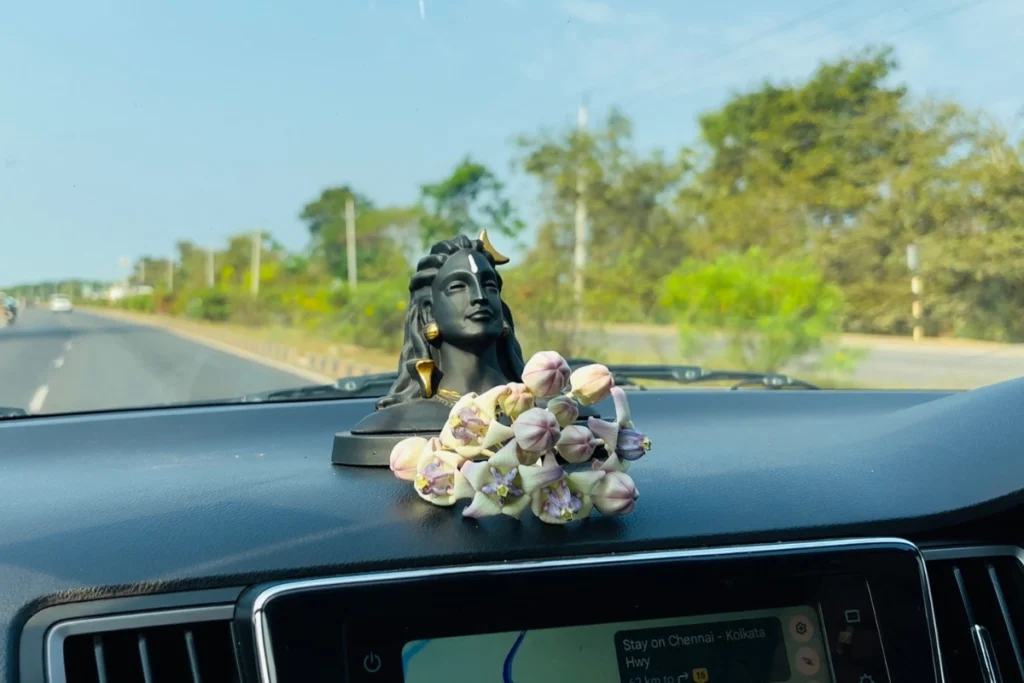 A view through a car windshield showing a rural highway with lush green trees on both sides and a small Lord Shiva idol on the dashboard.