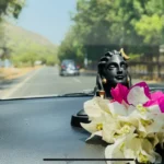 A close-up shot of a small black idol of Lord Shiva adorned with fresh pink and white flowers, placed on a car dashboard with a blurred highway visible through the windshield.