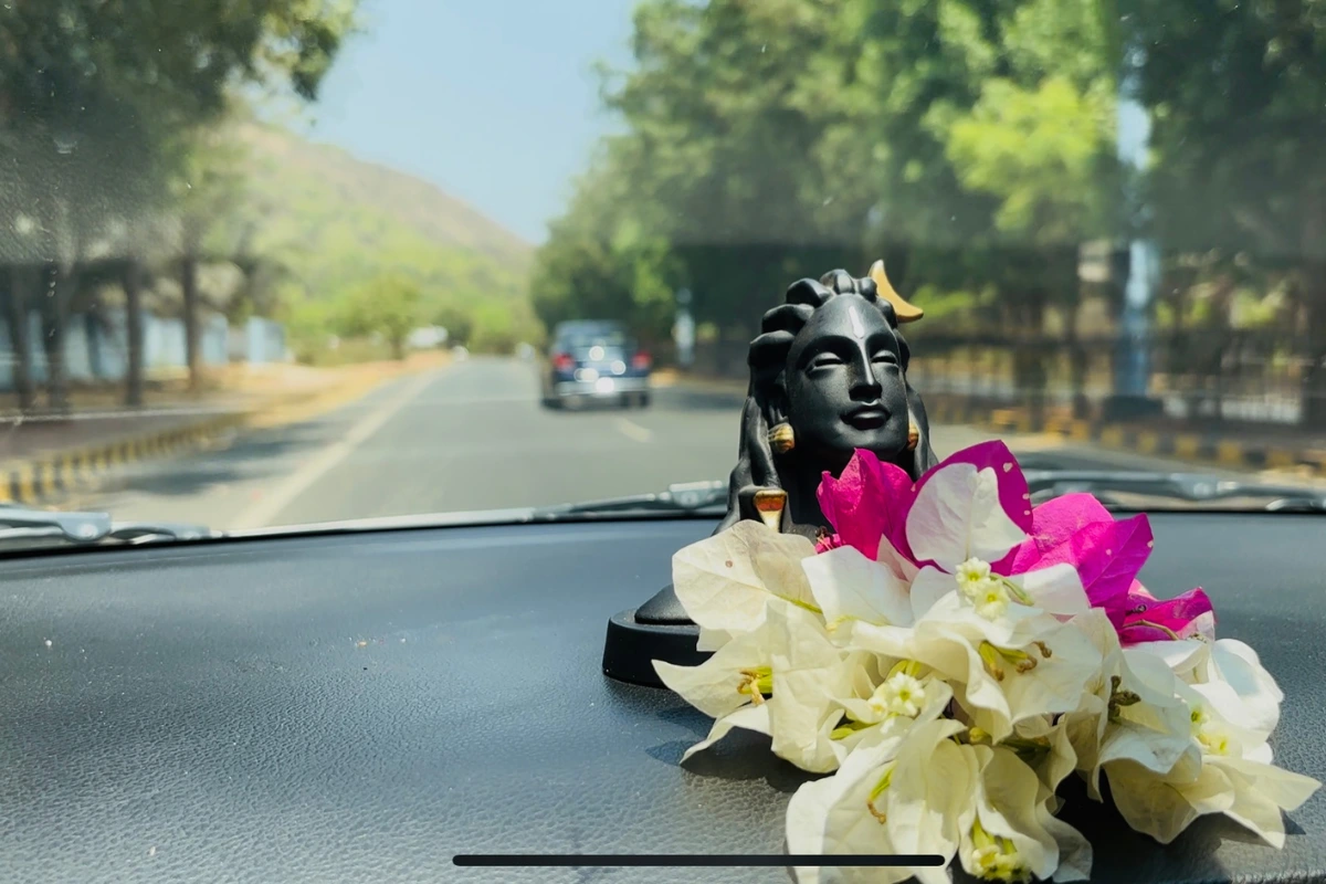 A close-up shot of a small black idol of Lord Shiva adorned with fresh pink and white flowers, placed on a car dashboard with a blurred highway visible through the windshield.