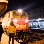 A side-angle shot of a long sleeper train illuminated under station lights at a busy terminal in the evening.