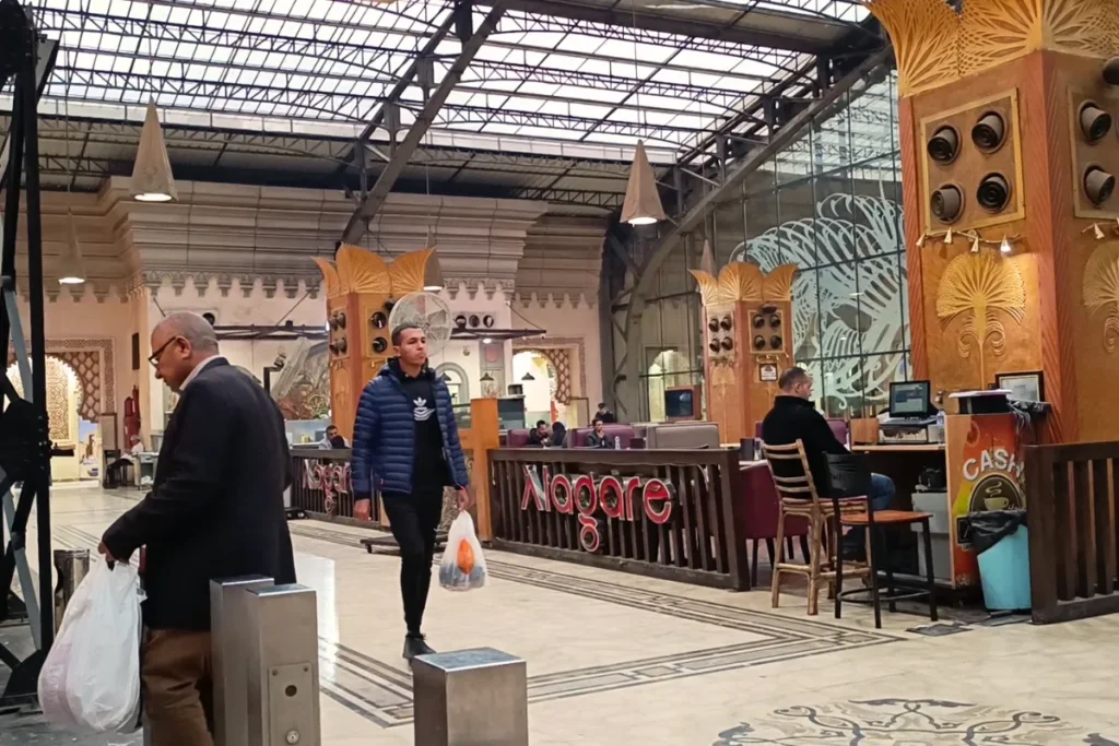 An interior view of a spacious cafe inside a historic train station terminal with arches, where people are walking by and a customer sits at a counter stool.
