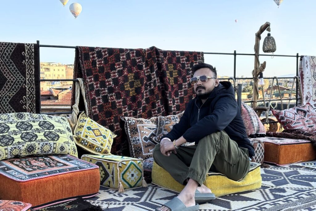Akash wearing sunglasses, a hoodie, and a dark jacket sits cross-legged on a yellow cushion on an outdoor rooftop terrace, posing for the camera with hot air balloons in the distant background.