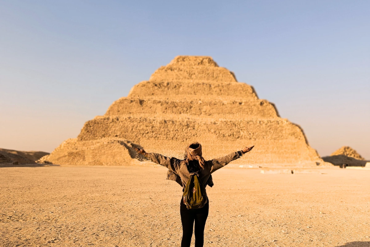 Sumana with a headscarf spreads their arms wide in front of the massive, tiered stone structure of the Step Pyramid under a clear blue sky.
