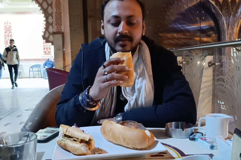 A close-up of Akash sitting at a table inside a station cafe, holding and enjoying a wrapped shawarma sandwich.
