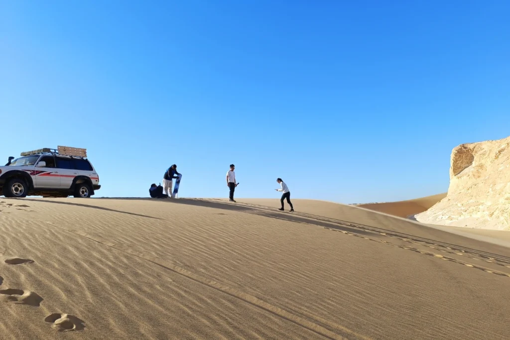 A green 4x4 vehicle parked at the base of a high sand dune as a person prepares to sandboard down.