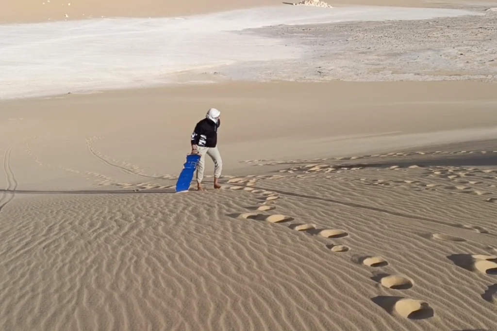Akash walking with a sandboard walking up a large sand dune in the desert.