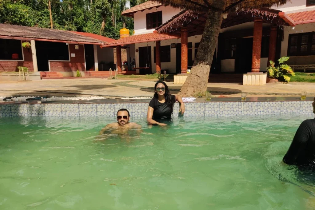 South India- Chikmagalur (2) Akash & Sumana enjoying themselves in a clear outdoor swimming pool with a resort building and trees in the background.