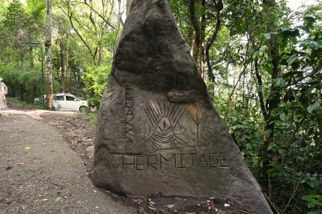 South India- Edakkel Cave 2 A large, triangular grey rock signpost engraved with the words "EDAKKAL HERMITAGE" and line-art symbols, standing next to a paved pathway with a white car and a dense green forest in the background.