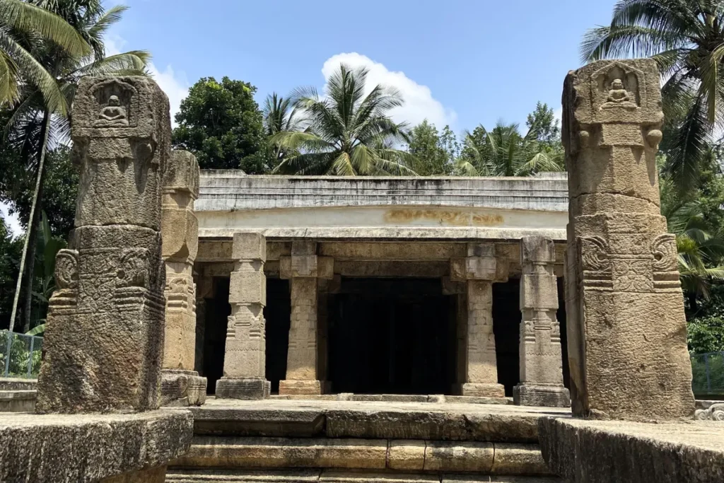 South India- Jain Temple 1 (1) The weathered stone entrance pillars and facade of an ancient Jain temple surrounded by grass and trees.
