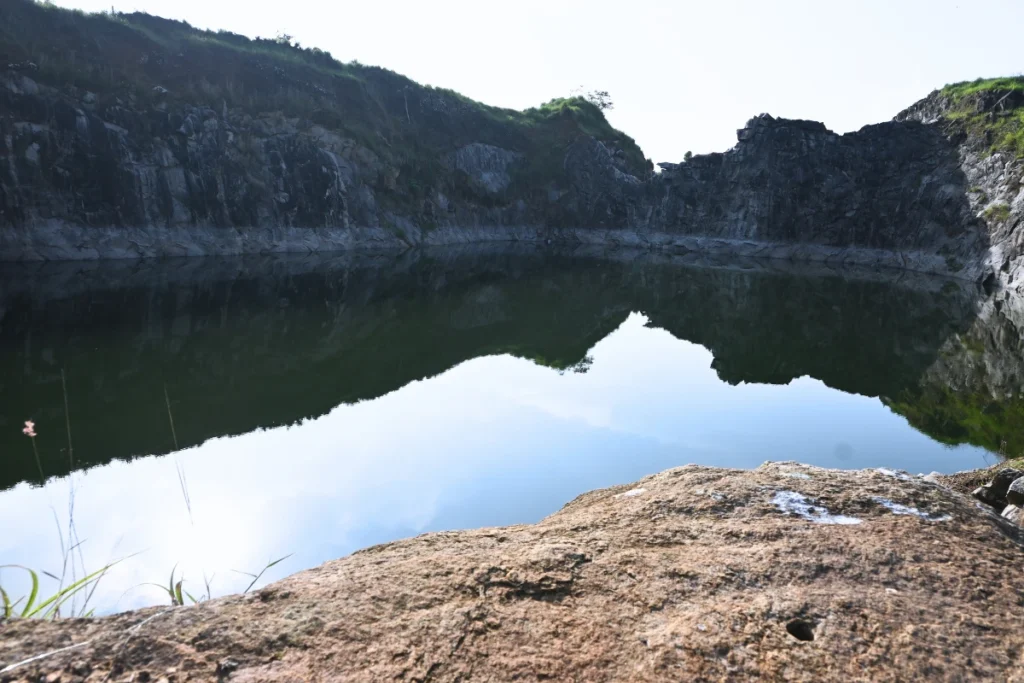 South India- Manjarappa lake A wide-angle landscape shot of the scenic Manjappada Quarry in Wayanad, Kerala. The image features a large, calm emerald-green water body pooled at the bottom of a deep-cut stone quarry. High, dark granite rock walls wrap around the pool, reflecting clearly onto the still water surface under a bright sky. In the immediate foreground, a rugged, sunlit boulder fills the lower frame.