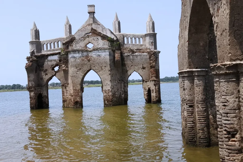 South India- Shettihelli Church The ruins of the gothic-style Shettihalli Rosary Church standing partially submerged in calm blue water under a bright sky.