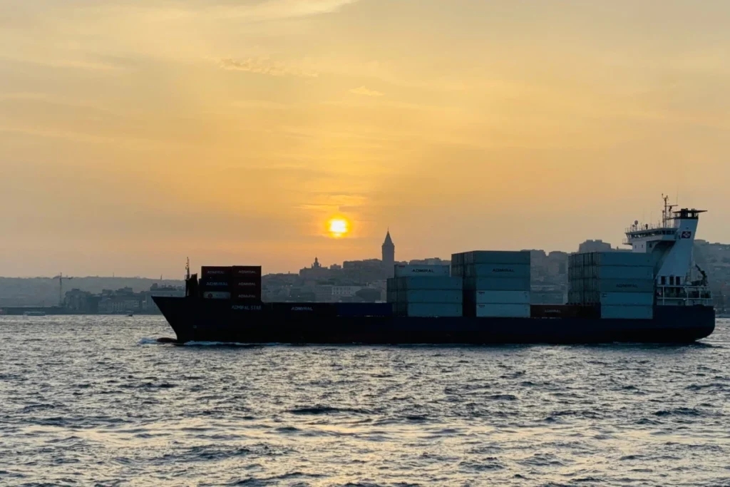 A silhouette of a large container cargo ship passing in front of a distant city skyline and Galata Tower during a golden sunset at the Bosphorus.