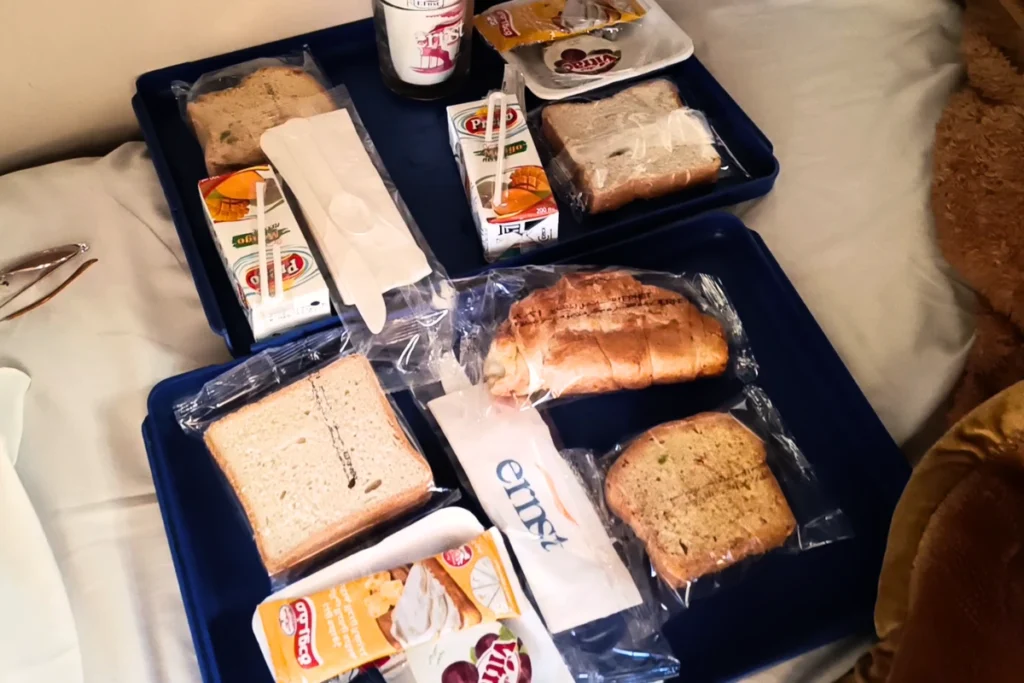 A packaged breakfast box laid open on a tray, featuring a croissant, cheese slices, bread rolls, and juice.