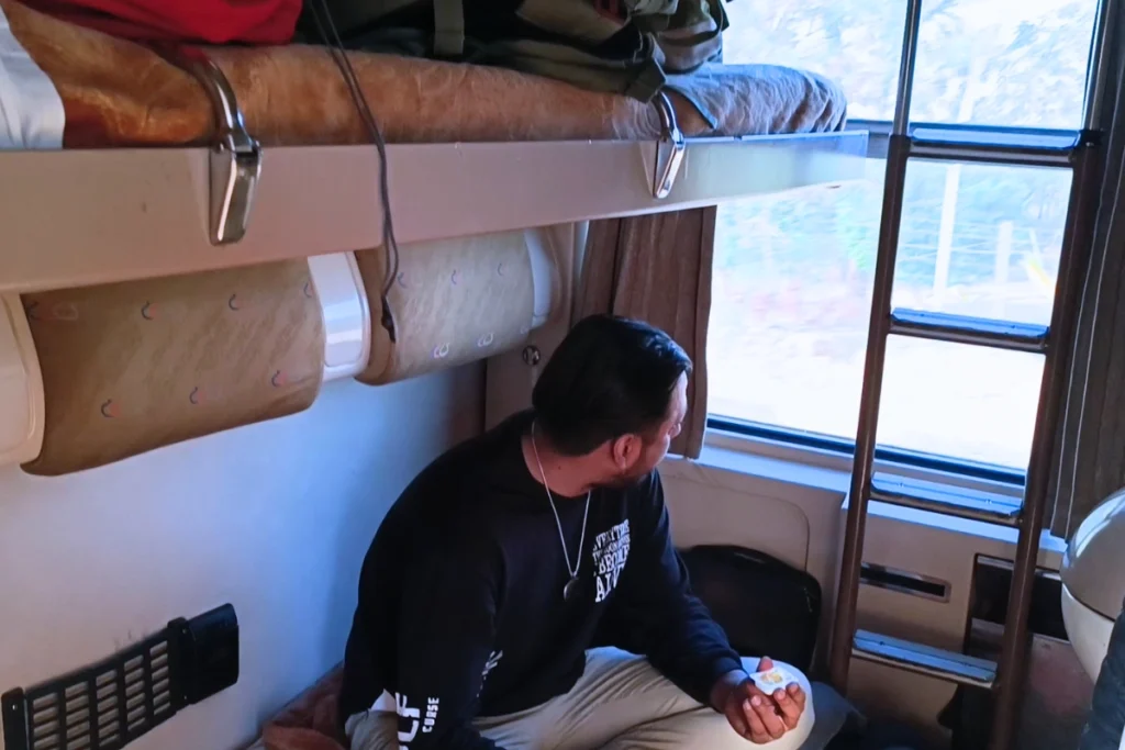 Akash sitting on the lower bunk of a compact train sleeper compartment, with the upper bunk folded down and a window looking out.