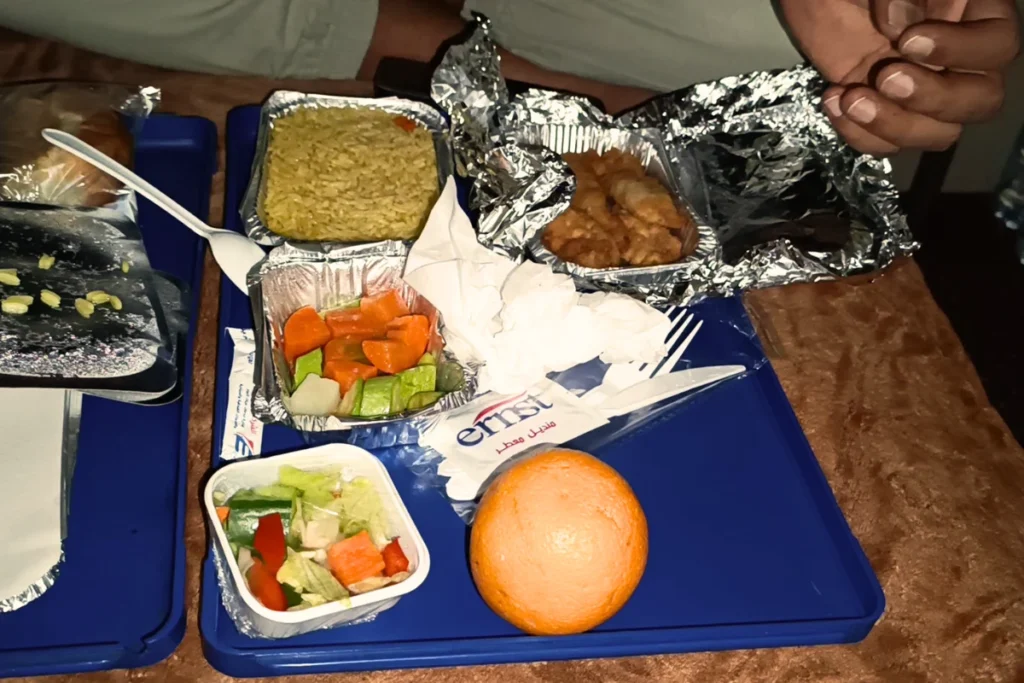 A flight-style meal tray on a small airplane-like table, containing chicken, rice, bread, orange slices, and small juice boxes.