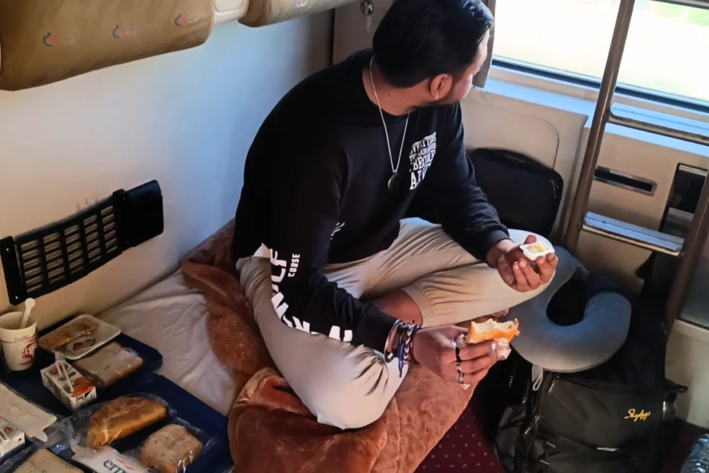 Akash sitting inside a train cabin, preparing a meal on a small fold-down table next to a window.