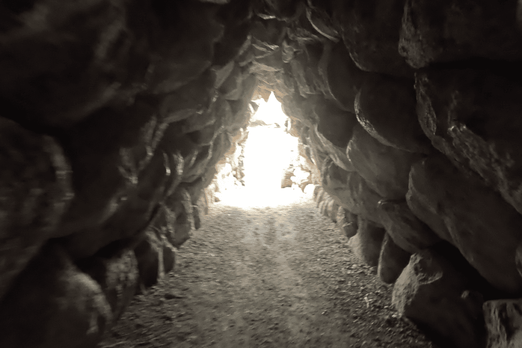 The interior view of a dark, corbelled stone tunnel leading toward a bright, sunlit exit.