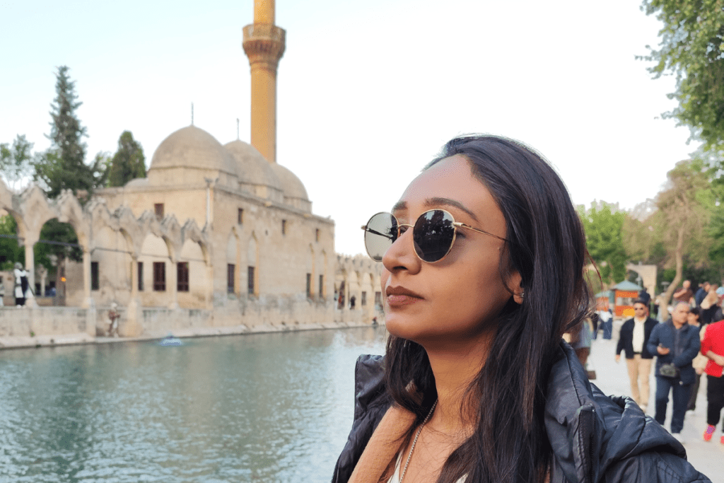 Turkey- Balekligol 1 Sumana wearing sunglasses poses in front of a sprawling, beautifully detailed ancient stone complex.