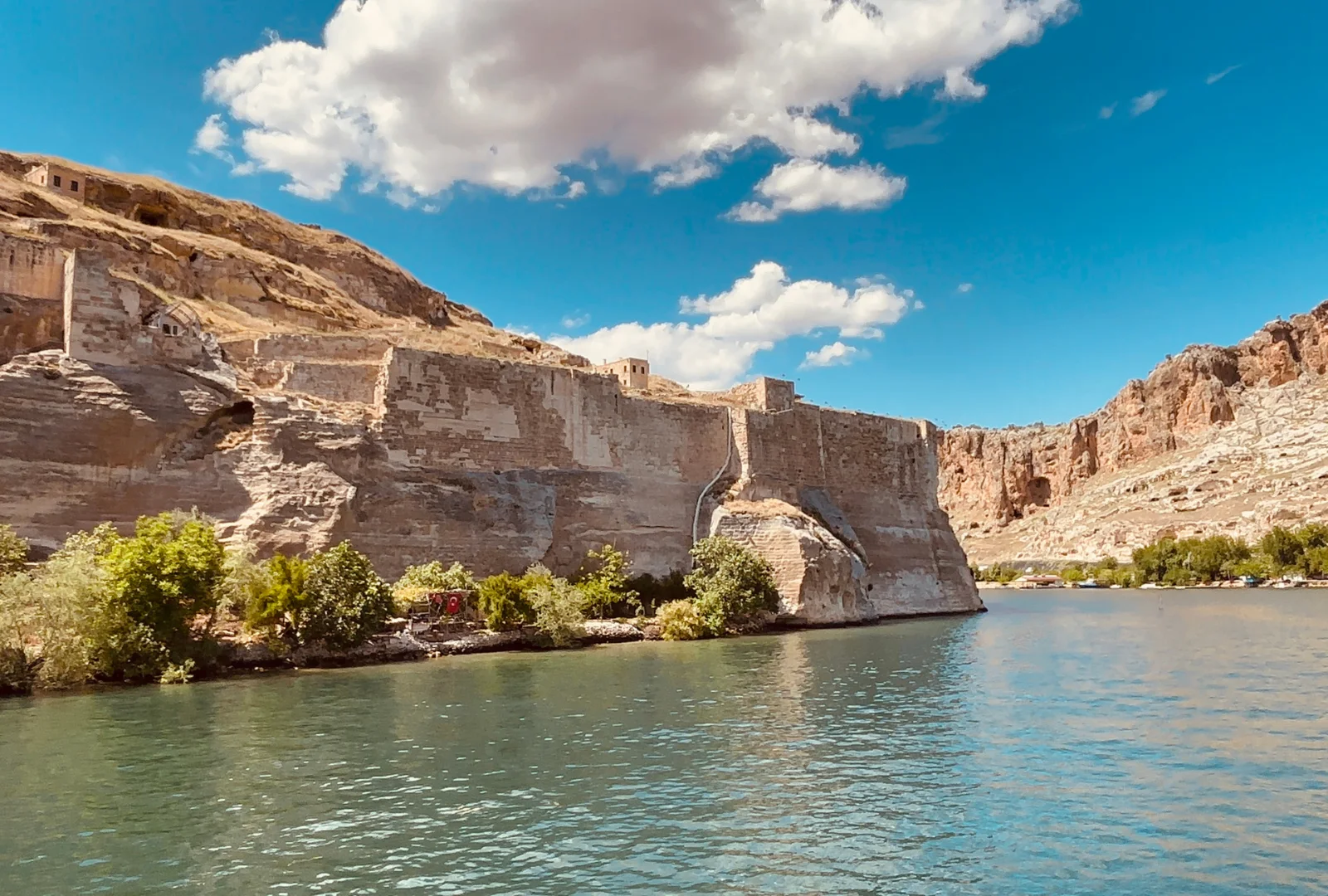 A horizontal landscape view showing a massive ancient stone fortress wall rising from the banks of a blue river under a bright sky.